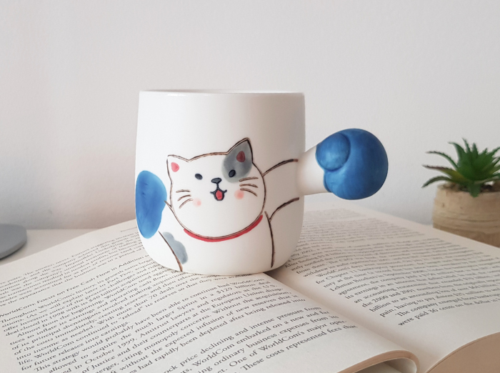 BOXING CAT MUG. A funny gift for coworkers. White mug with a cat drawn on it and as a holder it has the arm of the cat extended, with a blue glove, as if it was boxing. Rabbit is looking happy.